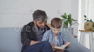 关爱的父亲正在教他的小儿子好奇的学龄前儿童阅读。 他们坐在沙发上看书